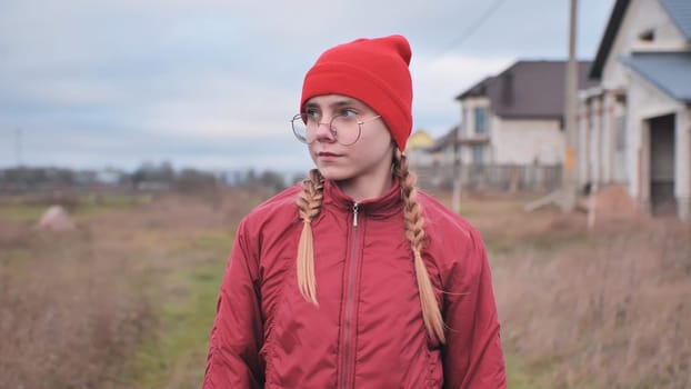 Portrait of a teenage girl wearing a red beanie, jacket, and glasses, with braided hair, standing in a field with houses in the background on an overcast day