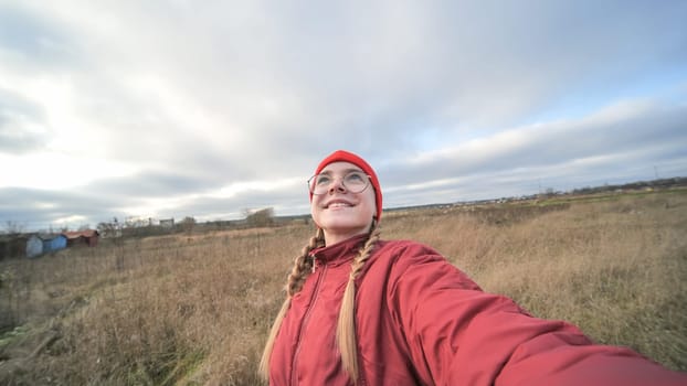 Smiling young woman with red winter clothes and glasses taking selfie in a countryside field, enjoying cloudy sky