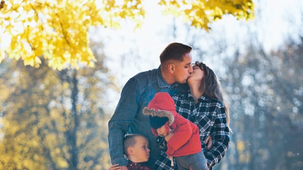 Loving parents embracing, sharing kiss while holding young children during golden autumn afternoon in scenic park setting