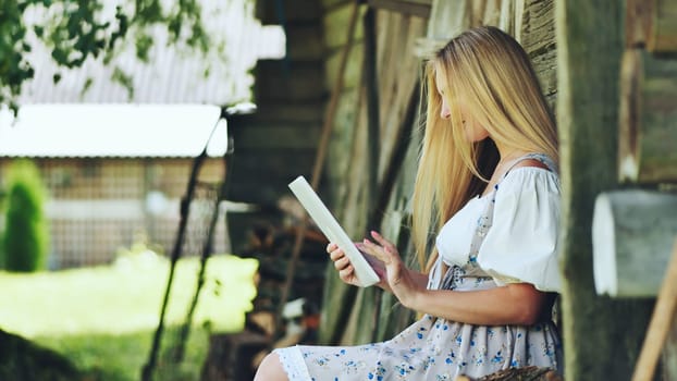 Blonde woman in a traditional dress uses a tablet while sitting next to a rustic wooden structure, blending technology with rural life
