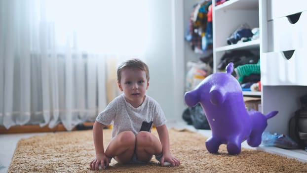 Little boy having fun in his bedroom sitting on a carpet and playing with an inflatable purple toy animal, next to a white wardrobe
