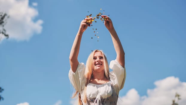 Slavic woman throwing gold confetti in the air, celebrating against a bright blue sky with fluffy white clouds