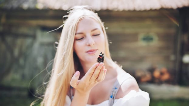 Young blonde woman gently holding a butterfly on her hand, with closed eyes and peaceful expression, in front of a rustic wooden house