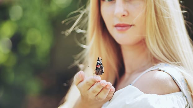 Blonde woman holding butterfly on her hand in green summer garden, enjoying nature and wildlife