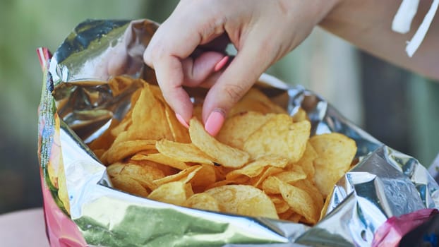 Female hand reaching into crisp foil bag, selecting salty potato chip with pink polished nails