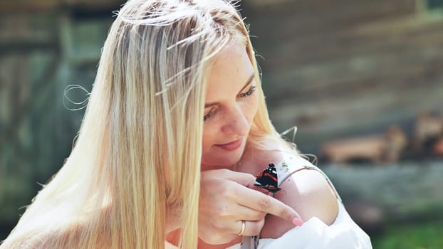 Blond woman with freckles gently touching butterfly standing on her shoulder with index finger