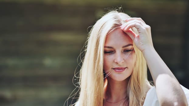 Blonde female caressing hair, bathed in warm sunlight near weathered wooden surface, expressing soft, relaxed demeanor