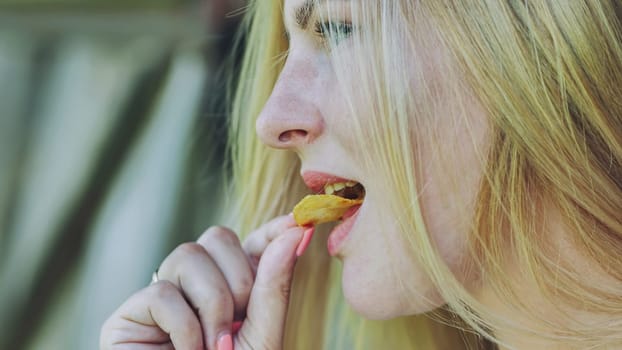 Close up profile of a cheerful blond woman savoring a crunchy potato chip, enjoying the salty, tasty snack on a sunny day outdoors