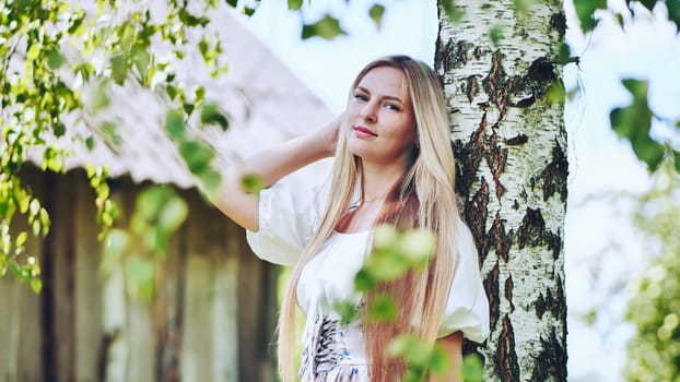 Blond woman in traditional slavic attire standing near birch, rural setting capturing cultural heritage and pastoral scenery
