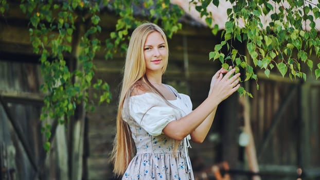 Young blonde woman wearing traditional slavic dress gently touching birch leaves in a peaceful countryside setting