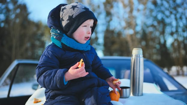 Toddler enjoying a snack on the car hood, surrounded by snow covered trees during a joyful winter family trip in cozy warm clothes