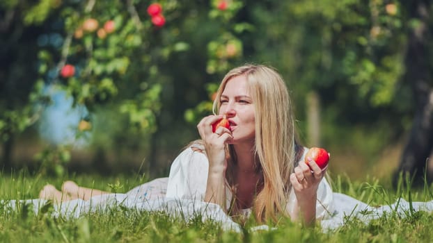 Blond woman relaxing, biting crisp red apple while reclining on white blanket amid lush green apple orchard rows