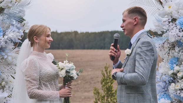 Loving groom reading handwritten vows, speaking into microphone, gazing adoringly at bride during rustic outdoor wedding ceremony