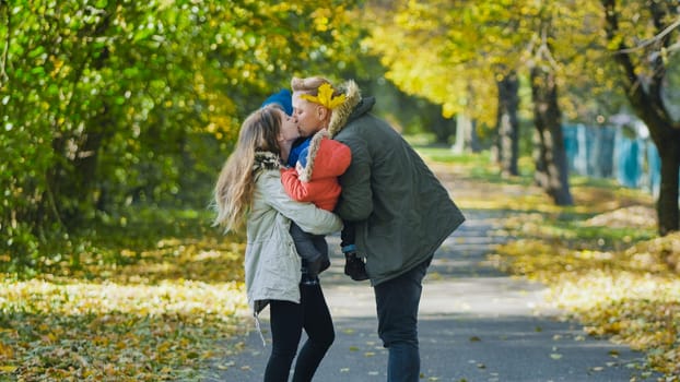 Parents kissing each other while holding their children in a park full of colorful autumn leaves