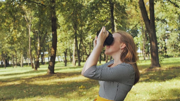 Young woman wearing virtual reality headset, experiencing immersive digital world amid green park landscape under bright sunlight