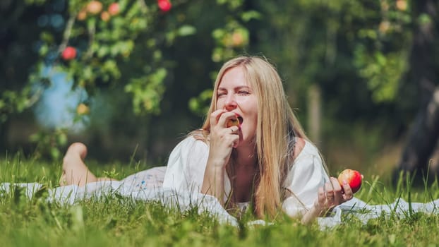 Young blond woman lying on blanket on grass eating apple in orchard, enjoying healthy snack on sunny summer day