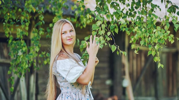 Young woman with long blond hair wearing traditional slavic dress gently touching birch leaves in countryside setting, celebrating her heritage