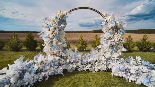 Beautiful wedding arch adorned with white and light blue flowers, set in a field under a partly cloudy sky, creates a picturesque ceremony backdrop