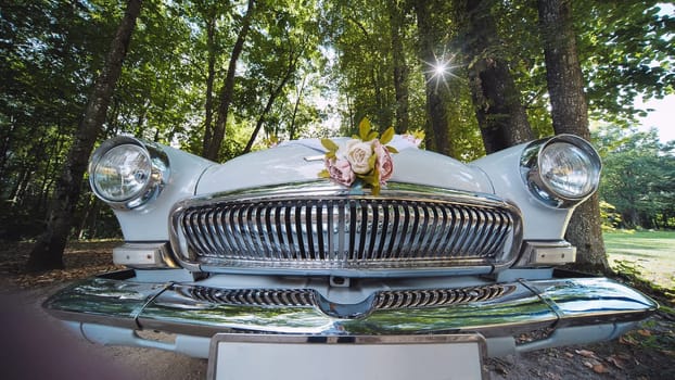 Vintage white car adorned with floral bouquet, parked near trees at wedding venue, symbolizing romantic celebration