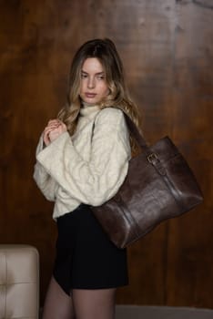 A girl in a warm sweater, a black short skirt, nylon tights is posing with a brown leather bag