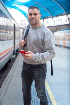 man in a gray sweatshirt standing at a train station, looking at his red smartphone. His relaxed expression reflects modern connectivity and urban travel looking at camera.