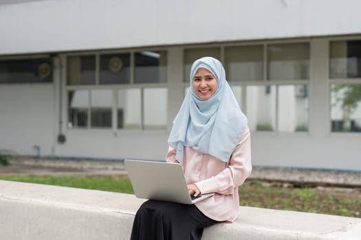 A smiling Muslim businesswoman in a hijab, comfortably working on her laptop in a peaceful outdoor environment, showcasing her professionalism.