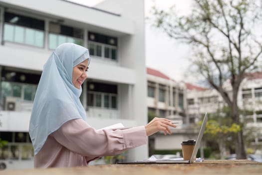 A cheerful Muslim businesswoman engaged in her work on a laptop, enjoying a coffee break in a vibrant outdoor setting.