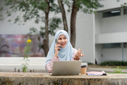 An enthusiastic Muslim businesswoman in a hijab engaged in a phone call, showcasing her vibrant work life in an outdoor setting.