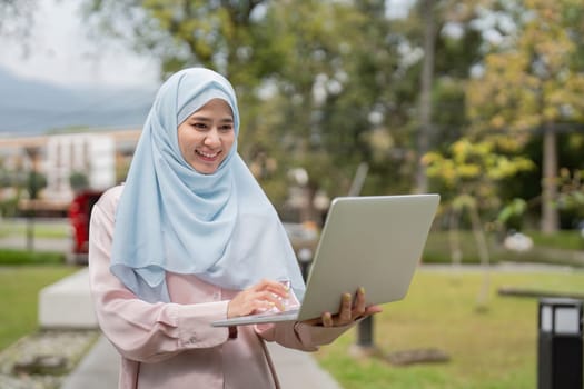 A happy Muslim businesswoman in a hijab, excitedly holding her laptop in a vibrant outdoor setting, reflecting her enthusiasm for work.