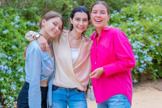 Three young women smiling and embracing each other while posing in a lush green park. They wear stylish casual outfits, expressing happiness, friendship, and joy in a natural outdoor setting.