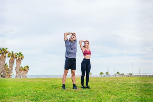 Young man and woman in sportswear stretching their arms in a green park near the coast. They are preparing for a workout, surrounded by palm trees and an open sky with fresh air.