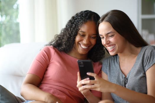 Happy interracial roommates using cell phone and laughing sitting on a couch at home