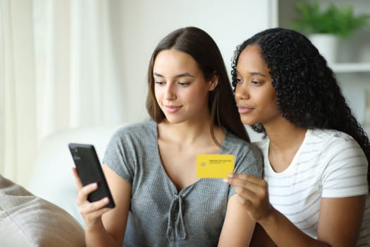 Interracial serious friends at home buying online with credit card and smart phone sitting on a sofa