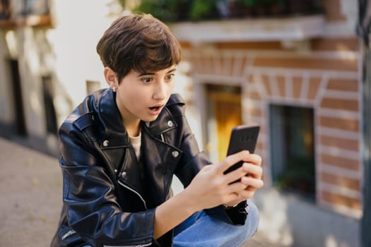 A youthful individual sporting a fashionable short haircut, and a stylish leather jacket is expressing a sense of astonishment as they gaze intently at their smartphone while enjoying the outdoors