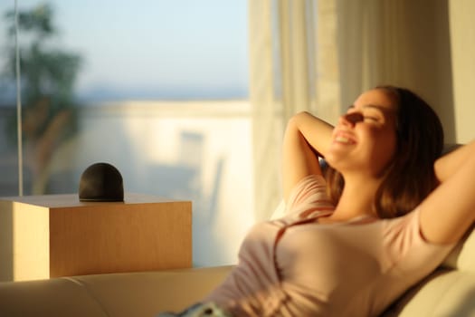 Woman relaxing at home at sunset with smart speaker sitting on a couch 