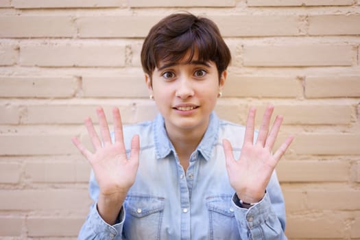 A young woman with a contemporary short haircut radiates vibrancy, expressing friendliness with her hands in a warm gesture against a textured wall that adds character to the scene