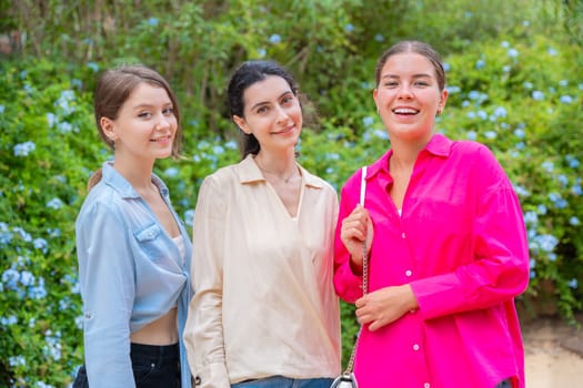 Three young women smiling and embracing each other while posing in a lush green park. They wear stylish casual outfits, expressing happiness, friendship, and joy in a natural outdoor setting.