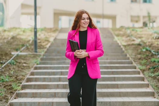 A confident businesswoman in a chic pink blazer ascends outdoor steps with a stylish notebook, embodying professionalism and grace, showcasing her determination to succeed in her career