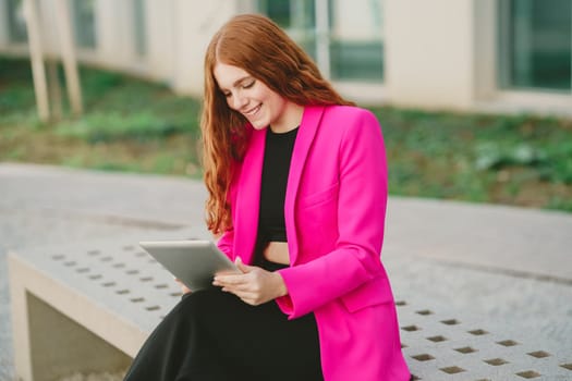 A stylish and fashionable woman dressed in a vibrant pink blazer is enjoying her time as she uses her tablet while seated outside in a modern and inviting setting, fully engaged in her activities