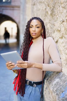 In a charming stone alley, a stylish young woman with braided hair stands confidently, engaged with her tablet, symbolizing modern living and the embrace of technology in an urban setting