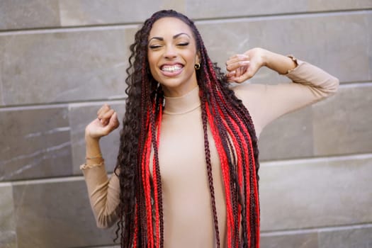 A vibrant young woman with long braided hair creatively styled in striking red and classic black, radiating joy as she smiles joyfully while posing outdoors against a beautifully textured wall
