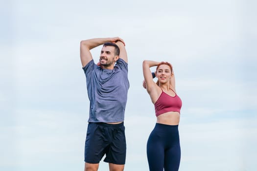 A fit man and woman stretching their arms before a workout in an outdoor setting. They wear sportswear and prepare their muscles against a bright sky, showing motivation and a healthy lifestyle.