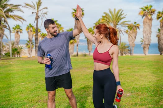 Smiling young man and woman celebrating after exercising outdoors. They are wearing sportswear and enjoying a joyful moment together in a green park with palm trees near the sea.