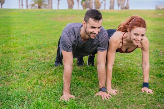 Smiling young man and woman doing push-ups together in a green park near the coast. Both are wearing sportswear and look motivated and energetic. The background features palm trees and nature.