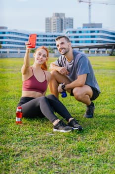 Smiling young man and woman sitting on the grass, taking a selfie after exercising. They are wearing sportswear and holding water bottles, enjoying a relaxed moment in an urban park.