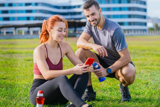 A young fit couple in sportswear relaxing after exercise in a green park. The woman is sitting on the grass using her smartphone, while the man crouches beside her with a water bottle, both smiling.