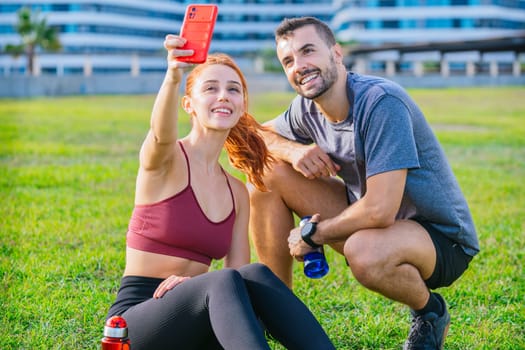 Smiling young man and woman sitting on the grass, taking a selfie after exercising. They are wearing sportswear and holding water bottles, enjoying a relaxed moment in an urban park.