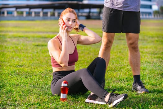 young athletic woman sitting on the grass in a park after exercise, talking on her smartphone. She wears sportswear and has a water bottle nearby. A man's legs are visible standing beside her.