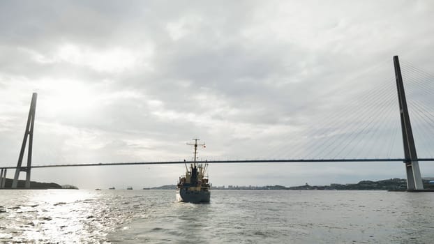 Cargo ship sailing beneath the russian bridge in vladivostok, russia, while clouds drift across the sky on a tranquil day
