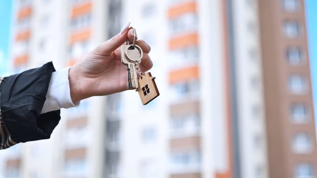 Real estate agent holding house keys featuring wooden home keychain, blurred residential complex background